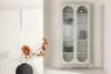 A tall, narrow beige display cabinet with glass doors and arched tops, standing in a minimalist room with a white wall and a vase with dried flowers. Voir les images du produit