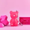 A pink bear-shaped keychain with a heart on its chest is displayed against a pink background. To the left are geometric pink heart shapes, and to the right is a pink rectangular sign with 'XOXO' written on it. Podgląd zdjęcia produktu