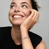 A woman with freckles smiles and looks up, resting her chin on her hand. She is wearing a black top and a silver bracelet with a heart-shaped charm. Anteprima dell’immagine prodotto