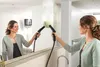 A woman is cleaning a mirror with a steam cleaner, with her reflection also visible. Anteprima dell’immagine prodotto