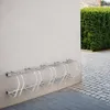 A metal bicycle rack mounted on a wall next to a paved area and a hedge. Náhled obrázku produktu