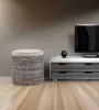 A woven storage ottoman with a cushioned top sits on a wooden floor next to a modern media console with a television. Podgląd zdjęcia produktu