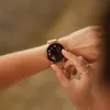 A close-up shot of a woman's wrist wearing a rose gold smartwatch with a floral watch face. Her finger is touching the screen. Anteprima dell’immagine prodotto