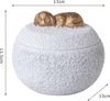 A white, textured round container with a lid, decorated with two gold-colored objects on top. Podgląd zdjęcia produktu