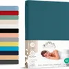 A teal fitted sheet is shown folded, with a stack of other colored fitted sheets to the left and packaging with a sleeping woman and product information to the right. Produktbild-Vorschau