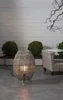A woven lamp with a light bulb inside sits on a concrete floor next to a wicker chair and a small table. Behind the lamp are two round green bushes, and a white wooden wall is in the background. A bare branch hangs from the upper right corner. Produktbild-Vorschau