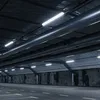 An interior view of a large, industrial-looking space, possibly a parking garage or warehouse, with prominent ductwork and lighting fixtures overhead. The walls appear to be rough-hewn concrete, and the floor is asphalt with painted lane markings. Produktbild-Vorschau
