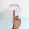 A hand is pressing the test button on a white smoke detector mounted on a white ceiling. Produktbild-Vorschau