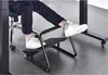 A person's legs and feet are shown resting on a black footrest under a desk. Náhľad obrázku produktu