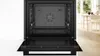 An open black built-in oven is shown in a well-lit room with a dining area in the background. Produktbild-Vorschau