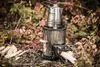 A camping stove with a coffee pot on top, set in an outdoor environment with fallen leaves and firewood. Produktbild-Vorschau