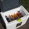 A cooler box filled with various bottles of drinks, placed outdoors on a paved surface with grass in the background. Produktbild-Vorschau