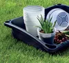 A black plastic tray filled with gardening supplies, including stacked white plastic pots, a small potted plant, soil, and bulbs, resting on green grass. Produktbild-Vorschau