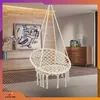 A white macrame hanging chair is shown in a living room setting, with a wooden wall and dresser in the background. Produktbild-Vorschau