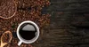 A close-up overhead view of a white coffee cup filled with black coffee, surrounded by roasted coffee beans on a dark wooden surface. A wooden spoon filled with coffee beans is also visible. Podgląd zdjęcia produktu