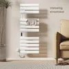 A white electric towel warmer is mounted on a light beige wall next to a beige sofa. A plant is visible on the left side of the image, and a rug is on the floor. Produktbild-Vorschau