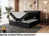 A bedroom scene featuring a black upholstered bed with its storage compartment open, showcasing the lifting mechanism. A large potted plant and decorative items are also visible. Produktbild-Vorschau