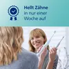 A woman is smiling and holding two electric toothbrushes, looking into a mirror. Produktbild-Vorschau