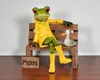 A decorative frog figurine wearing a yellow raincoat and boots, sitting on a wooden bench next to a small duck figurine. A wooden sign with 'MOIN' is placed in front of the bench. Produktbild-Vorschau