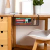 A black metal hanging basket filled with books and pens is attached to the underside of a wooden desk. A white chair is partially visible in the foreground. Produktbild-Vorschau