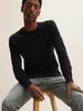 A man wearing a black sweater and grey jeans sits on a stool against a light background. Produktbild-Vorschau