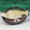 A novelty singing fish mounted on a wooden-like plaque, set against a green wooden plank background. Produktbild-Vorschau