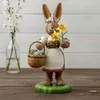 Ein fröhlicher Osterhase mit großen Ohren hält eine gelbe Blume in der rechten Hand und trägt in der linken einen Korb voller bunter Ostereier während er auf einem grünen Sockel steht und eine weiße Schürze mit blauen Details trägt Produktbild-Vorschau
