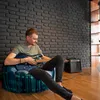 A man is sitting on a bean bag chair and playing an electric guitar in a room with a brick wall. Voir les images du produit