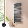 A modern anthracite electric towel radiator mounted on a light pink wall next to a window with greenery visible outside. A small white side table with a plant is also present. Náhled obrázku produktu