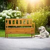 A wooden garden bench with a storage compartment is placed on a paved patio in a garden setting, with a golden retriever dog lying on the patio next to it. Lush green foliage and white flowers form the background, with sunlight filtering through the trees. Náhled obrázku produktu