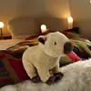 A plush capybara toy sits on a bed with a colorful blanket and fluffy rug. Produktbild-Vorschau