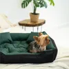 A small dog is resting on a green and black dog bed, with a plant and a wooden table in the background. Voir les images du produit