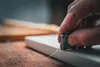 A close-up shot of a hand using a tool to scrape a white surface, with wood in the background. Produktbild-Vorschau