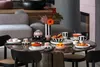 A dining table set with Villeroy & Boch Memphis tableware, including a vase, bowls, plates, and cups, along with croissants and fruit. The setting is in a modern living space with a sofa in the background. Voir les images du produit