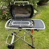A portable grill is set up on a grassy lawn with some greenery in the background. Produktbild-Vorschau