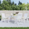 A white outdoor dining set with cushions is arranged on a patio, surrounded by lush greenery and a wooden fence. Produktbild-Vorschau