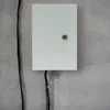 A white electrical junction box mounted on a concrete wall with black cables running alongside and into it. Náhled obrázku produktu