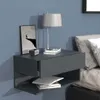 A modern gray wall-mounted nightstand with a drawer and open shelf, placed next to a bed with gray and white bedding. A lamp, a framed picture, and a clock are on top of the nightstand. Books are placed in the open shelf. Produktbild-Vorschau