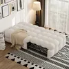 A beige tufted sofa bed with a throw blanket is placed on a patterned rug in a room with framed pictures on the wall and a floor lamp. Produktbild-Vorschau