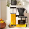 A yellow Moccacoffee maker sits on a white countertop, with a bowl of fruit to the left and a plant in a pot in the background. Podgląd zdjęcia produktu