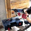 A person is using a Bosch hammer drill to drill into a wooden beam, with the drill's case and accessories visible. Náhľad obrázku produktu