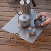 A person's hand is reaching for a cup on a small metal camping table with a kettle and teapot. Náhled obrázku produktu