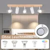 A ceiling light fixture with five adjustable spotlights mounted on a wooden bar, shown above kitchen cabinets. Below the fixture, there are images of different light bulbs. Produktbild-Vorschau