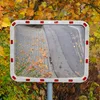 A traffic mirror with red and white reflectors on its frame, reflecting a paved path and a road with fallen autumn leaves. The mirror is surrounded by trees with yellow and orange leaves. Podgląd zdjęcia produktu