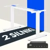 A white electric standing desk frame is shown from a diagonal angle, with a blue backdrop and the text '2 SILNIKI' in large white letters on a dark background strip across the foreground. A black control panel with a digital display is in the bottom right  Podgląd zdjęcia produktu
