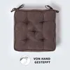A brown tufted seat cushion with ties, shown from a top-down perspective against a white background. A circular graphic with a hand sewing and text is present in the bottom right corner. Náhled obrázku produktu