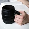 A person's hand holds a black grenade-shaped mug in front of an open book. Voir les images du produit