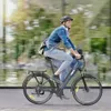 A man is riding an electric bicycle on a street with a blurred background of buildings and trees. Produktbild-Vorschau