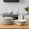 A set of white porcelain plates and bowls are neatly stacked on a wooden table in a kitchen setting. In the background, a stovetop and backsplash are visible, along with a glass carafe of water and a glass. Produktbild-Vorschau