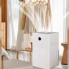 A white dehumidifier is placed on the floor in front of a clothing rack with several garments hanging on it. The dehumidifier has a power button with a circular indicator light. Produktbild-Vorschau